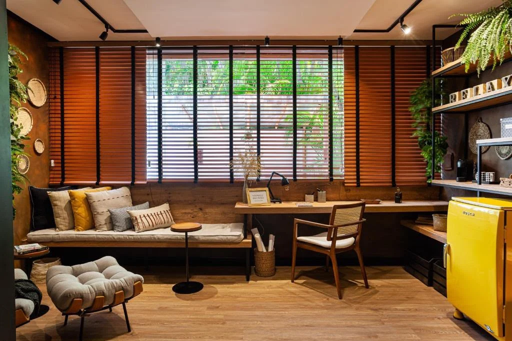 Espaço com bancada de trabalho, banco com almofadas, plantas e persianas de lâminas em tom de madeira
