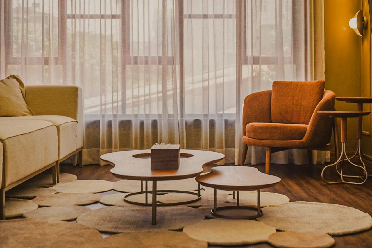 Sala com cortinas translúcidas, sofá claro, poltrona terracota e tapete de formas orgânicas