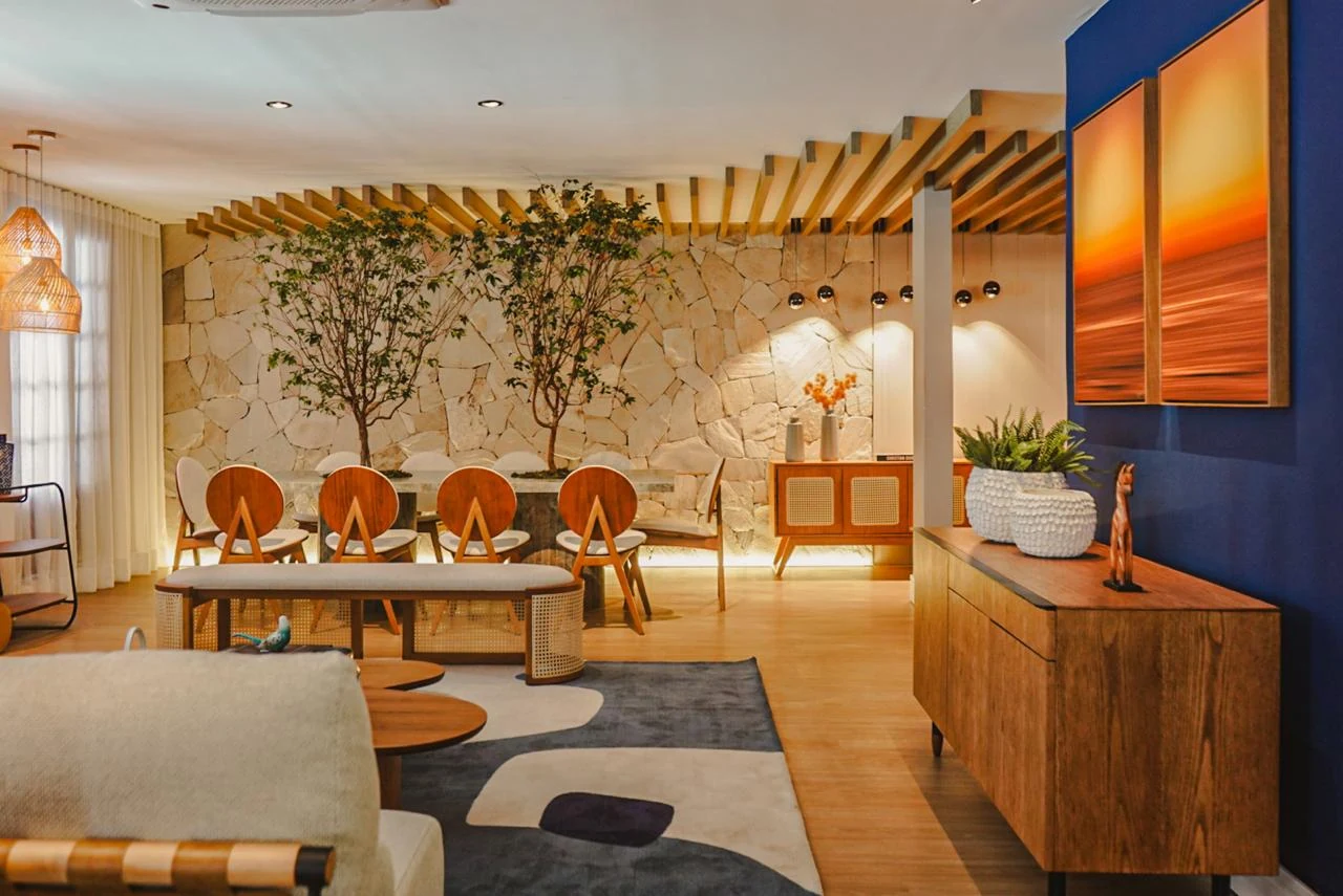 Sala integrada com cortinas claras, parede de pedra, árvores junto à mesa de jantar e mobiliário de madeira